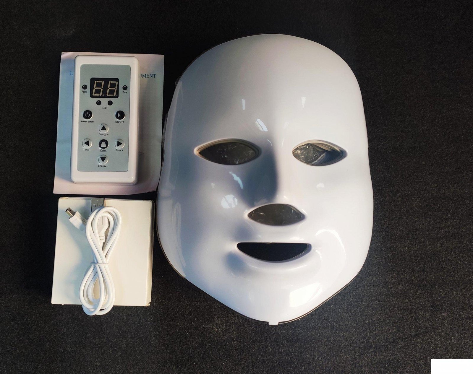 White LED face mask with its controller, charging cable, and manual laid out on a dark surface